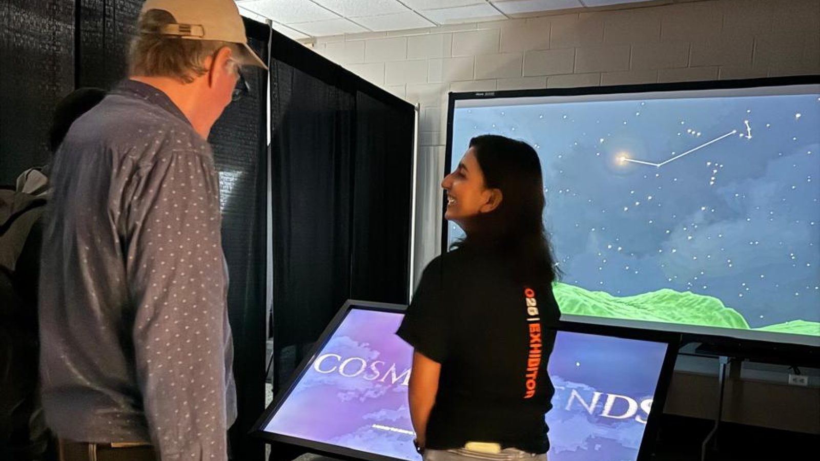 Neha talking with a visitor beside the kiosk and the big sky screen